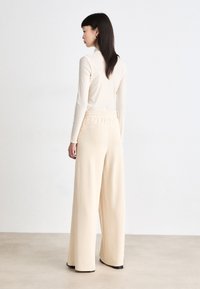 Beige langermet topp med høy krage, kombinert med vide kremfargede bukser med elastisk midje, glatt tekstur og minimalistisk design.