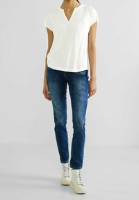 White short-sleeve top with a V-neck design, paired with blue denim jeans. White high-top sneakers complete the outfit.