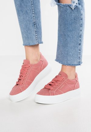 Różowe sneakersy na platformie z sznurowadłami, noszone z jasnoniebieskimi dżinsami z postrzępionym dołem na białym tle.