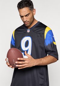 Σκούρα μαύρη φανέλα ποδοσφαίρου με μπλε και κίτρινες λεπτομέρειες, με τον αριθμό 9. Ένα αρσενικό πρόσωπο κρατάει ένα καφέ ποδόσφαιρο μπροστά του.