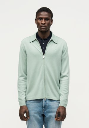 Muž nosiaci svetlozelený kardigán na zips cez čierne polo tričko a svetlomodré džínsy, stojaci pred jednoliatym bielym pozadím.