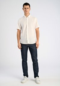 Vit kortärmad skjorta med knappar i linne, kombinerad med mörkblå slim-fit jeans och vita sneakers mot en neutral bakgrund.