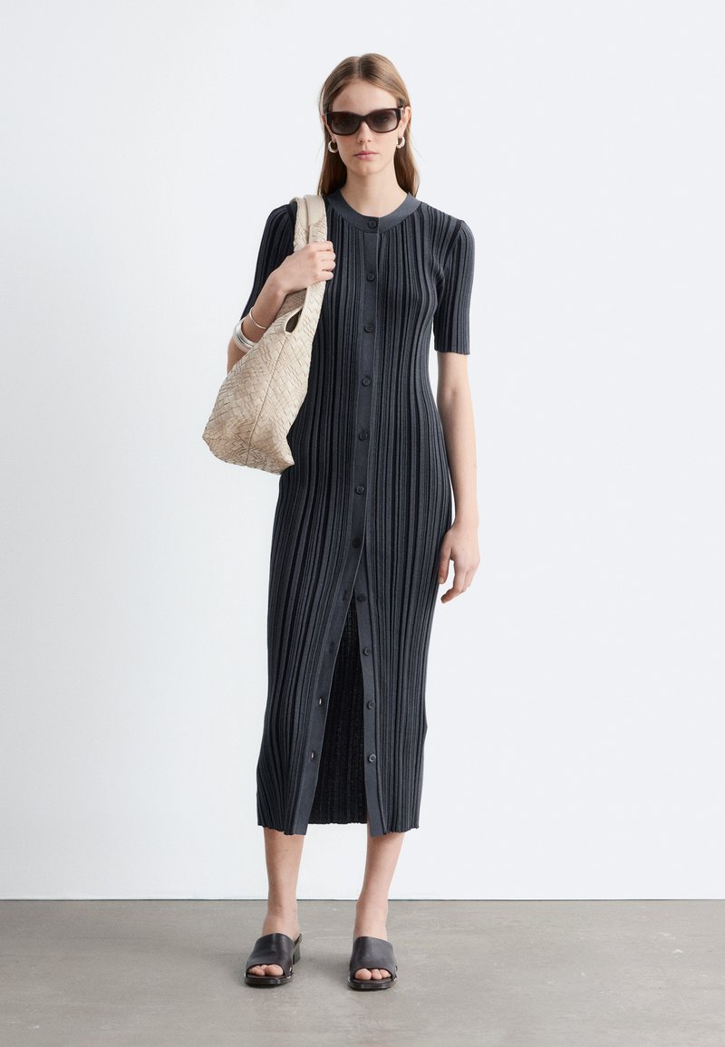 Femme portant une robe midi noire côtelée avec boutons sur le devant, manches courtes, sandales slide noires, lunettes de soleil, bracelets argentés et portant un sac tissé beige.