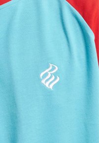 Trumpos rankovės marškiniai, padalinti į turkio ir raudonos spalvos audinį. Turkiškoje dalyje yra baltas siuvinėtas logotipas su banguotomis linijomis.