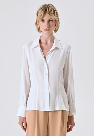 Camicia bianca con colletto classico, maniche lunghe e vita aderente. Tessuto liscio con sottili linee verticali.