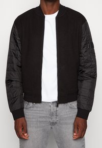 Zwarte bomberjack met een fleece lichaam en gewatteerde nylon mouwen. Heeft een ritssluiting aan de voorkant en aansluitende manchetten. Gedragen over een wit T-shirt.