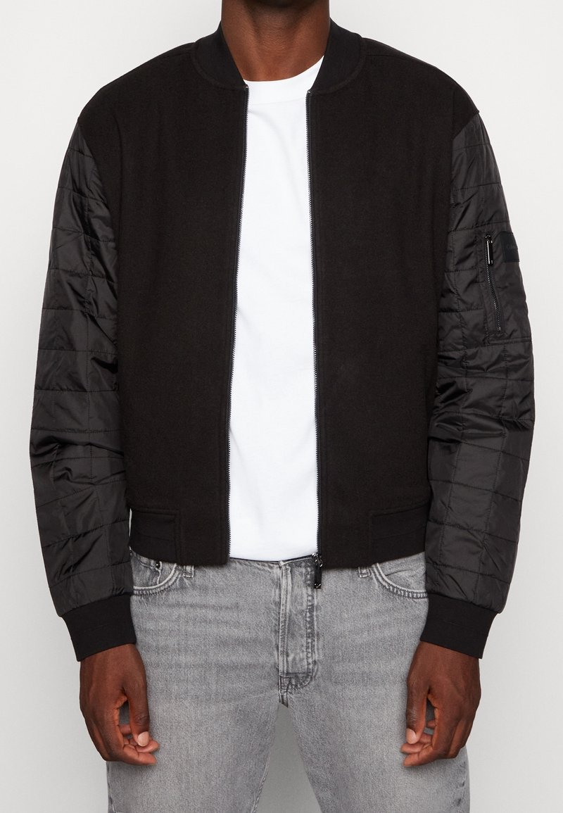 Zwarte bomberjack met een fleece lichaam en gewatteerde nylon mouwen. Heeft een ritssluiting aan de voorkant en aansluitende manchetten. Gedragen over een wit T-shirt.