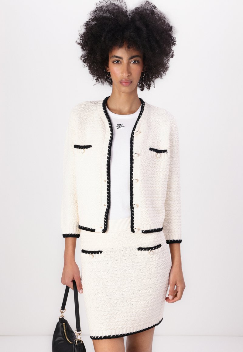 Biały kardigan z fakturą, z czarnymi wykończeniami, złotymi guzikami i dwiema przednimi kieszeniami. W komplecie ze spódnicą w pasującym stylu, eksponującą dopasowany krój.