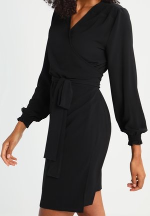 Mujer vistiendo un vestido cruzado ajustado negro con mangas largas abullonadas y un cinturón atado a la cintura, de pie contra un fondo claro y liso.