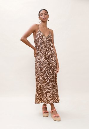 Mujer con vestido sin mangas marrón y blanco con estampado, tirantes delgados y sandalias plataforma coral, de pie sobre un fondo liso.