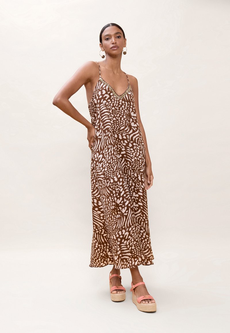 Nainen, jolla on päällään ruskeavalkokuvioinen hihaton mekko ohuilla olkaimilla ja korallinväriset platform-sandaalit, seisomassa yksiväristä taustaa vasten.