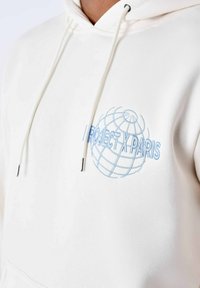 Hoodie bianco con un design ricamato di un globo blu e il testo "PROJECT X PARIS". Presenta una tasca frontale a marsupio e un cappuccio con cordino.
