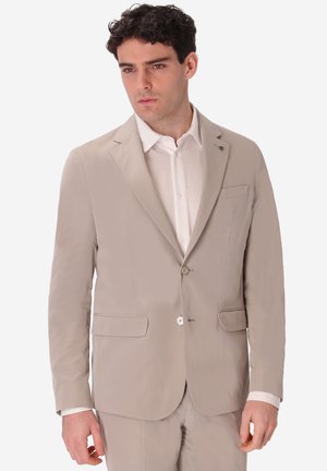 Uomo che indossa giacca beige e pantaloni coordinati sopra una camicia bianca, in piedi davanti a uno sfondo semplice.