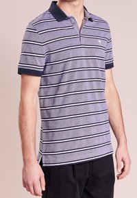 Pruhované polo tričko v odstínech šedé, s námořnicky modrým límcem a lemy rukávů, s malým logem na hrudi. Vyrobené z bavlněného materiálu.