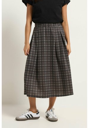 Femme portant une jupe plissée à carreaux foncés mi-longue, un haut noir à manches courtes et des baskets blanches à rayures noires, debout devant un fond blanc.