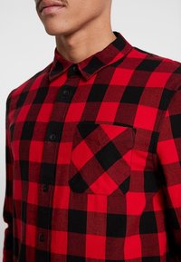 Camicia in flanella a quadri rossi e neri con colletto abbottonato, tasca frontale e bottoni neri. La trama del tessuto sembra tessuta.