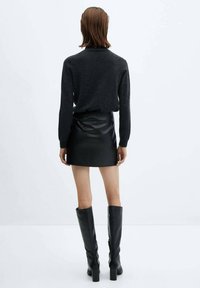 Pull noir à col roulé, mini-jupe noire ajustée en fausse cuir, et bottes noires à talons montant jusqu'aux genoux, vus de dos sur fond blanc.