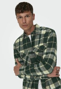 Camisa de franela a cuadros verdes y amarillos con dos bolsillos en el pecho, cierre de botones en la parte delantera y una textura única. El modelo está de pie con los brazos cruzados.