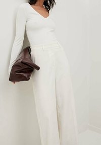 Top bianco a maniche lunghe con scollo a V, abbinato a pantaloni cream a vita alta e a gamba larga. Viene tenuta una borsa in pelle marrone spiegazzata.