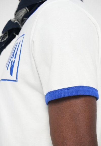 Bela majica s kratkimi rokavi in modrimi rebrastimi manšetami. Na sprednji strani je modra grafika. Tkanina se zdi mehka in gladka.