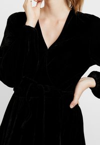 Žena v černých sametových šatech s dlouhými balónovými rukávy, svázanými v pase, pózuje s jednou rukou na bocích a druhou blízko tváře.