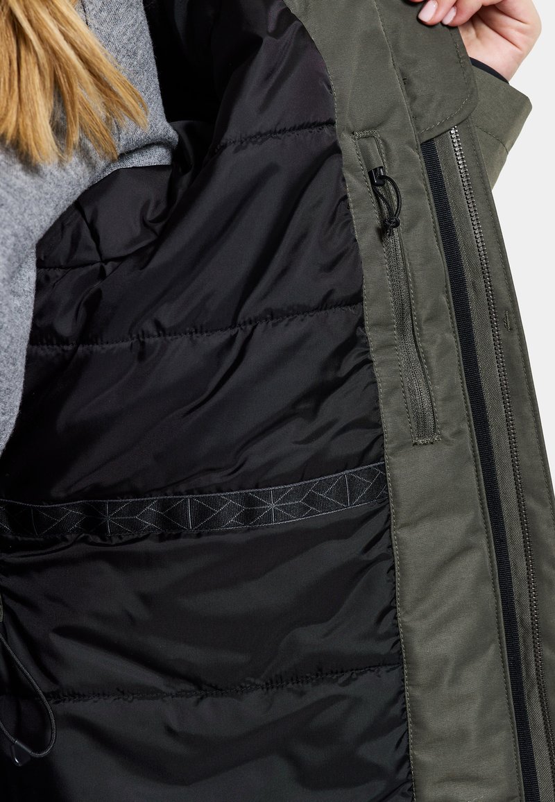 Grüne Außenjacke mit schwarzem Innenfutter, ausgestattet mit einer Reißverschlusstasche und elastischen Details. Das Material erscheint strapazierfähig mit einer glatten Textur.