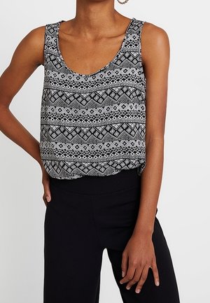 Mujer con un top sin mangas de patrón geométrico en blanco y negro y pantalones negros de cintura alta, posando con una mano en la cadera.