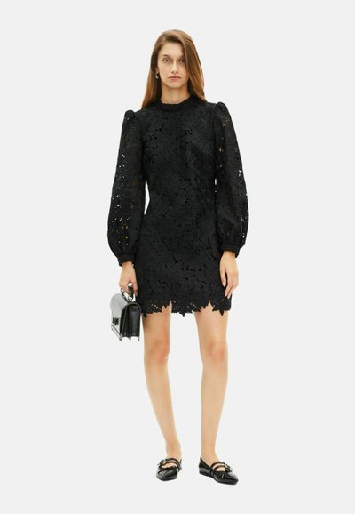 Frau in einem schwarzen, blumigen Spitzen-Minikleid mit langen Ärmeln, die eine schwarze Handtasche hält und schwarze spitze Ballerinas mit Schnallen-Trägern trägt.