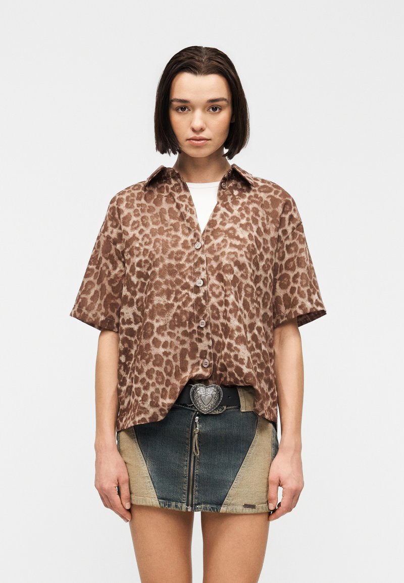 Νεαρή γυναίκα με κοντά σκούρα μαλλιά που φοράει καφέ μπλουζάκι με λεοπάρ μοτίβο και μίνι φούστα τζιν δίχρωμη με αγκράφα ζώνης σε σχήμα καρδιάς.