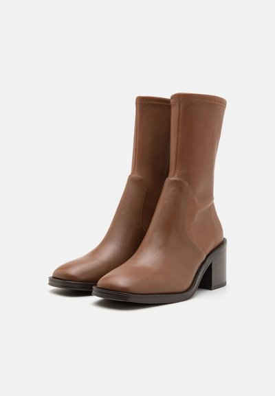 Bottes en cuir marron à mi-mollet au design épuré, avec un talon bloc et une texture lisse. Elles présentent une pointe légèrement arrondie et des coutures minimalistes.