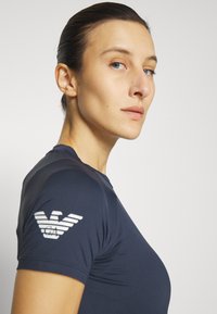 Marineblauw aansluitend t-shirt van rekbare stof, met een subtiele textuur en een witte logoprint op de linkerschouder.
