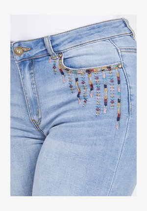 Svetlo modri kavbojki z zapletenimi večbarvnimi vezeninami ob sprednjem žepu, s čopiči, ki visijo spodaj.