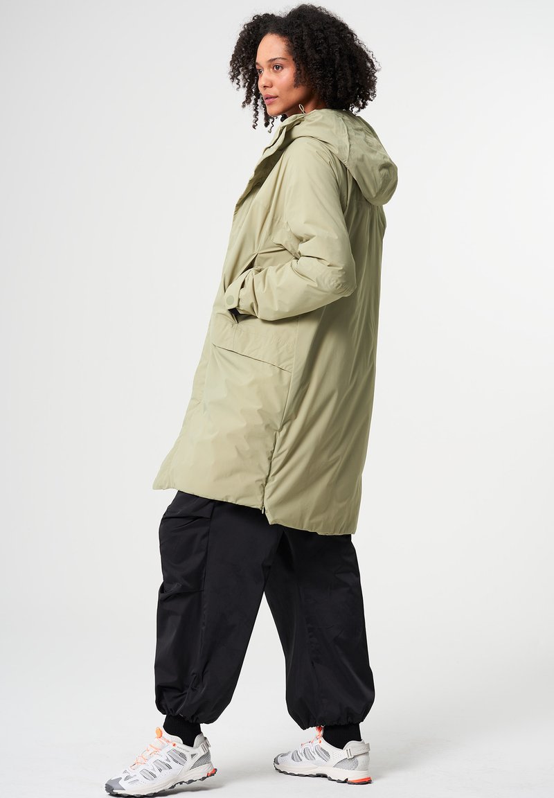 Helle olivgrüne wasserdichte Jacke mit Kapuze und Fronttaschen, kombiniert mit lockeren schwarzen Hosen und Sportschuhen. Glatte Textur.