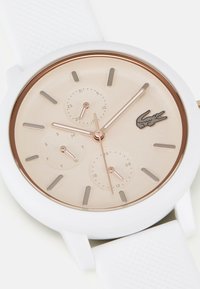 Reloj redondo con correa de silicona blanca, esfera beige y acentos en oro rosa. Presenta tres subesferas para la fecha, el día y la hora en formato de 24 horas.