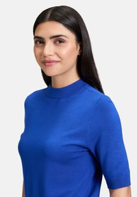 Maglietta a maniche corte in maglia blu con collo a giro, trama liscia e design aderente, indossata da un modello su uno sfondo neutro.