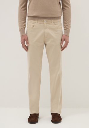 Uomo che indossa pantaloni beige a gamba dritta, mocassini marroni e un maglione beige a maniche lunghe, in piedi davanti a uno sfondo chiaro e uniforme.