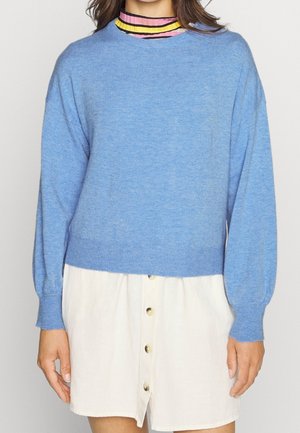 Femme portant un pull bleu clair par-dessus un col roulé rayé rose, jaune et noir, associé à une jupe blanche à boutons sur le devant.