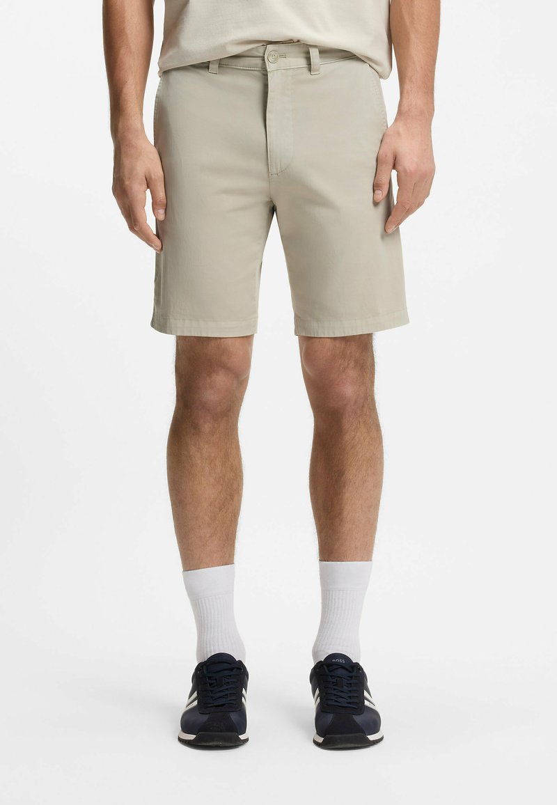 Homme portant un short beige jusqu'aux genoux, des chaussettes blanches montantes et des baskets noires, debout, les bras détendus le long du corps.