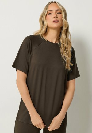 Femme aux longs cheveux blonds portant un t-shirt ample à manches courtes marron foncé, debout devant un fond clair uni.