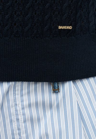 Μαύρο πουλόβερ με πλεκτό τελείωμα και μικρή χρυσή ετικέτα "SANDRO" πάνω από τη μέση μπλε και λευκής κάθετης ριγέ φούστας ή παντελονιού.