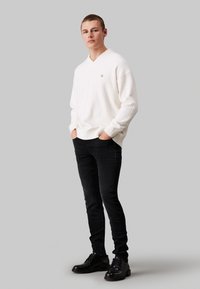 Suéter blanco de cuello en v con puños acanalados, combinado con jeans negros ajustados y botas negras con cordones. El suéter presenta un pequeño logo en el pecho.
