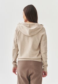 Bež hoodie s kapuljačom na vezanje, rebrastim manžetama i donjim rubom. Mekana tekstura i opušteni kroj, u kombinaciji s smešnim hlačama.