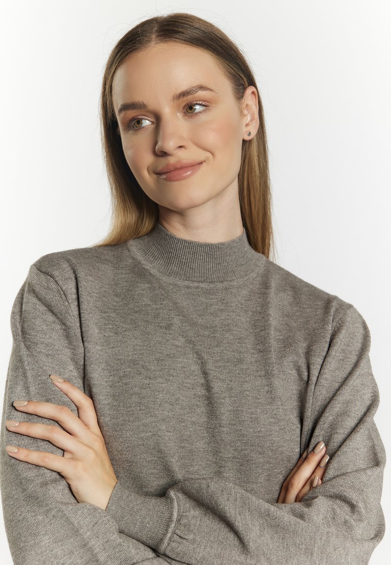 Junge Frau mit langen Haaren, die einen grauen Pullover trägt, mit verschränkten Armen, leicht zur Seite blickend und mit einem subtilen Lächeln.