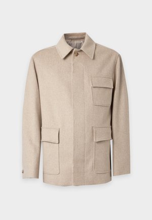 Mand iført beige jakke med fire lommer, lyseblå skjorte, mørke løse bukser og mørke sko, stående mod en ensfarvet hvid baggrund.