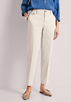 Kobieta ubrana w beżowe spodnie o prostych nogawkach, niebieską bluzkę z długim rękawem oraz taupeowe spiczaste baleriny, stojąca z jedną ręką w kieszeni.