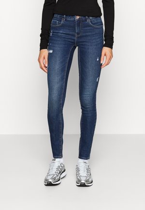 Jeans ajustados de mezclilla azul oscuro con parches desgastados y de cintura media. Combinados con zapatillas deportivas grises y negras.