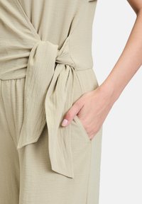 Lys beige jumpsuit med en front wrap detalje, bundet i livet. Har en lomme på siden og et blødt, tekstureret stof.