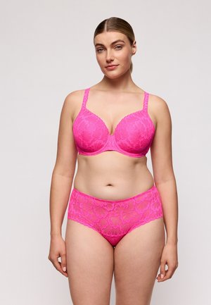 Vrouw met lichte huid en blond haar die een bijpassende felroze kanten beha en slip met hoge taille draagt, staand tegen een effen lichte achtergrond.