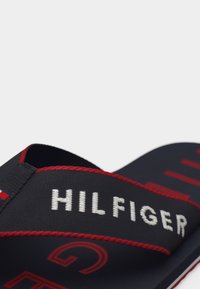 Ρουμπινί και κόκκινος λουρί σανδαλιού με λευκή κεντημένη επιγραφή "HILFIGER" και κόκκινη ραφή σε σανδάλι slip-on.