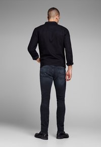 Man i svart jacka och mörka smala jeans, står med ryggen mot kameran med en hand i bakfickan mot en grå bakgrund.
